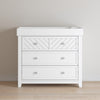 The Child Craft Atwood 3-Drawer Dresser in matte white features two top drawers with a chevron pattern and one larger plain drawer below, shown on a light wood floor against a beige wall.