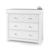 The Child Craft Atwood 3-Drawer Dresser in matte white has a chevron pattern on the top drawer and pink soap and lotion dispensers on the right side of its surface.