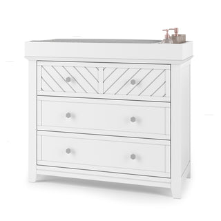 The Child Craft Atwood 3-Drawer Dresser in matte white has a chevron pattern on the top drawer and pink soap and lotion dispensers on the right side of its surface.