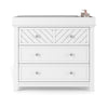 The Child Craft Atwood 3-Drawer Dresser in matte white features a chevron pattern on the top drawer, silver knobs, and is shown with a gray changing pad and baby supplies on top.