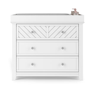 The Child Craft Atwood 3-Drawer Dresser in matte white features a chevron pattern on the top drawer, silver knobs, and is shown with a gray changing pad and baby supplies on top.