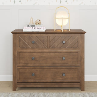 The Child Craft Atwood 3-Drawer Dresser in Cocoa Bean features geometric patterns and is topped with a lamp, books, and decor. It sits against a white paneled wall with subtle wallpaper above.