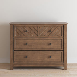 The Child Craft Atwood 3-Drawer Dresser in Cocoa Bean features a chevron pattern on the top drawer and black square knobs, set on a light wood floor against a plain white wall.