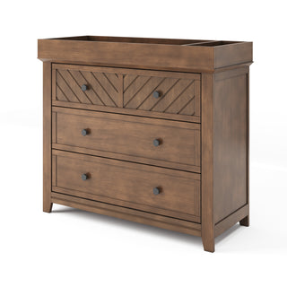 The Child Craft Atwood 3-Drawer Dresser in Cocoa Bean features three drawers with simple black knobs, a chevron-patterned top drawer, raised edge on top, and a warm brown finish.