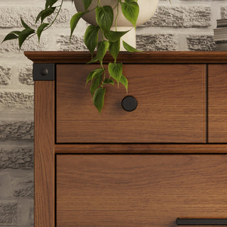 A close-up of the Child Craft Redmond 4 Drawer Chest in Coach Cherry shows its round black knob and wood grain beside a white brick wall, with a leafy green plant in a white pot resting on top of the dresser.