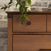 A close-up of the Child Craft Redmond 4 Drawer Chest in Coach Cherry shows its round black knob and wood grain beside a white brick wall, with a leafy green plant in a white pot resting on top of the dresser.
