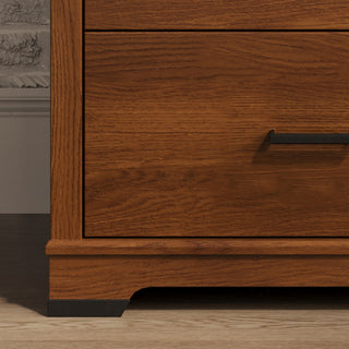 Close-up of the bottom corner of the Child Craft Redmond 4 Drawer Chest in Coach Cherry with a visible drawer and black handle, set on a light wood floor near a white wall—an ideal rustic bedroom furniture piece.