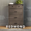 The Child Craft Redmond 4 Drawer Chest in Dapper Gray stands against a gray paneled wall, topped with a plant and books. It features spacious drawers, black metal pulls, and comes in two finishes.