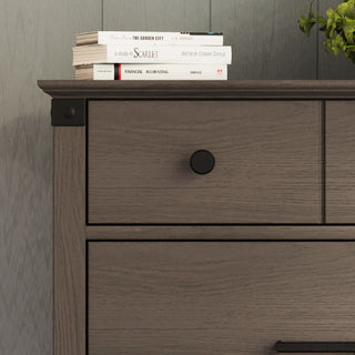 A close-up of the Child Craft Redmond 4 Drawer Chest in Dapper Gray, featuring two visible drawers with black knobs, a stack of books including The Scarlet and Arthur Conan Doyle on top, and a plant in the upper right corner.