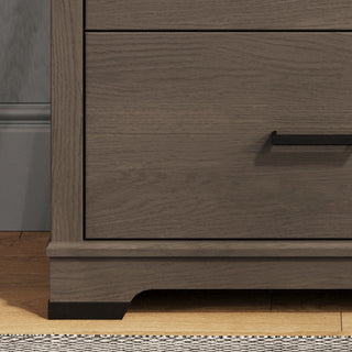 A close-up of the bottom corner of the Child Craft Redmond 4 Drawer Chest in Dapper Gray, featuring a black handle. The chest sits on a wood floor next to a gray wall and a rug with a striped edge.