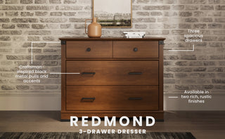 The Child Craft Redmond 3 Drawer Dresser in Coach Cherry stands against a light brick wall, featuring craftsman-style black metal pulls, spacious drawers, and a rich finish. A vase and framed picture rest on top.