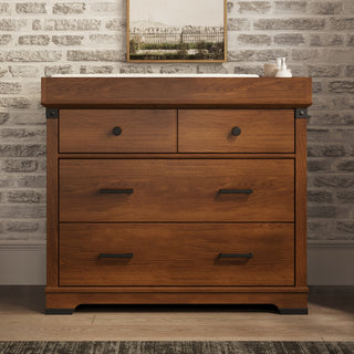The Child Craft Redmond 3 Drawer Dresser in Coach Cherry stands against a white brick wall, with a framed picture and small items placed on top. The dresser features two small upper drawers, a larger bottom drawer, and black handles.