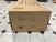 A large brown cardboard box with black text and symbols sits on a white tile floor with black diamond accents. The box displays shipping and product details for the Child Craft Redmond 3 Drawer Dresser in Coach Cherry.