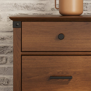 Close-up of the Child Craft Redmond 3 Drawer Dresser in Coach Cherry, featuring a round black knob on the top drawer and a black handle below, set against a white brick wall with a brown ceramic pitcher partly visible.
