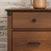 Close-up of the Child Craft Redmond 3 Drawer Dresser in Coach Cherry, featuring a round black knob on the top drawer and a black handle below, set against a white brick wall with a brown ceramic pitcher partly visible.