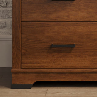 A close-up of the Child Craft Redmond 3 Drawer Dresser in Coach Cherry shows two drawers with black handles, standing on a light tile floor beside a gray brick wall.