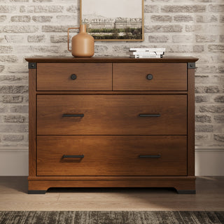 The Child Craft Redmond 3 Drawer Dresser in Coach Cherry stands against a light brick wall, topped with a tan vase, two stacked books, and a small framed picture hanging above.
