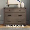 The Child Craft Redmond 3 Drawer Dresser in Dapper Gray has black metal pulls and craftsman-inspired style, making it perfect for a nursery. Two vases and a photo frame on top add a decorative touch.