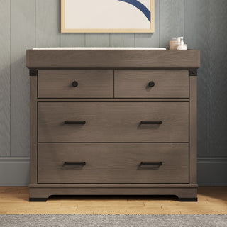 A Child Craft Redmond 3 Drawer Dresser in Dapper Gray, with black handles and toiletries on top, is placed against a paneled wall beneath a framed abstract artwork.
