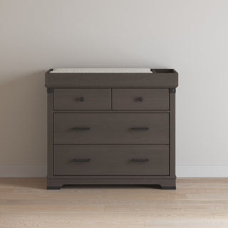A dark wood dresser with four drawers stands on a light wood floor against a pale wall, topped with the Child Craft F01792 Universal Changing Table Topper in Dapper Gray.