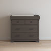 A Child Craft Redmond 3 Drawer Dresser in Dapper Gray stands against a plain light-colored wall on a wooden floor, topped with a changing pad.