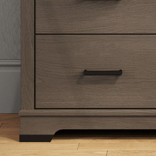 A close-up of the Child Craft Redmond 3 Drawer Dresser in Dapper Gray shows two drawers, each with a simple black handle. The dresser stands on a light wood floor beside a gray wall with white trim.