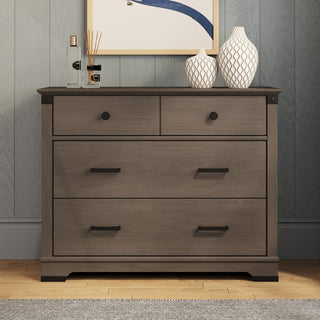 The Child Craft Redmond 3 Drawer Dresser in Dapper Gray features two small top drawers and a large bottom drawer, styled with vases and decor, and sits beneath artwork—a chic, practical addition to any nursery.