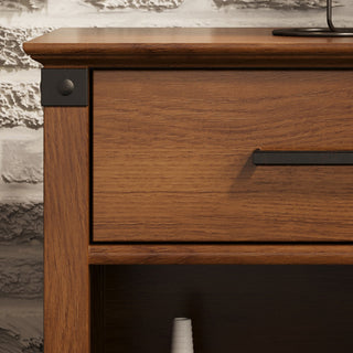 A close-up of the Child Craft Redmond Nightstand in Coach Cherry, featuring a drawer and open shelf with black metal handles and corner accents. A textured, painted brick wall serves as the background.