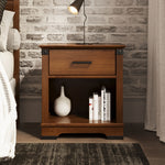 The Child Craft Redmond Nightstand in Coach Cherry stands by a white brick wall, topped with a black lamp and paired with a bed with a white blanket. Its open shelf holds a white vase and books.