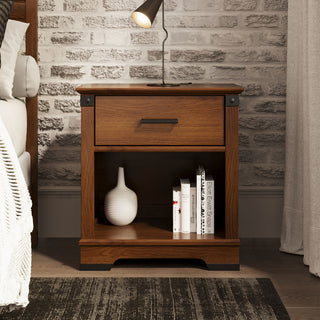 The Child Craft Redmond Nightstand in Coach Cherry stands by a white brick wall, topped with a black lamp and paired with a bed with a white blanket. Its open shelf holds a white vase and books.
