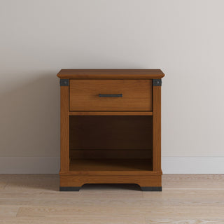 The Child Craft Redmond Nightstand in Coach Cherry features a single drawer with a metal handle, an open lower shelf, and minimal black accents. It stands on a light wood floor against a plain, light-colored wall.