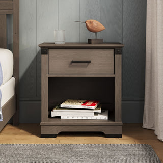 The Child Craft Redmond Nightstand in Dapper Gray features a drawer and open shelf with books. Topped with a glass of water and a wooden bird, it sits by the bed against a gray paneled wall.