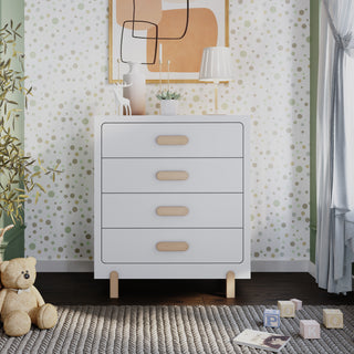 The Child Craft Park Heights 4-Drawer Chest in Matte White stands in the nursery with wooden handles, topped by a lamp, framed abstract art, and decor. Nearby, a teddy bear, plant, and alphabet blocks rest on a patterned rug against green dot wallpaper.