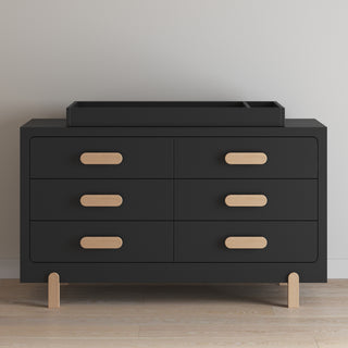 A sleek black six-drawer dresser with light wood handles and legs stands against a plain wall. On top sits the Child Craft Legacy Changing Table Topper in Caviar Black.