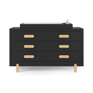 The Child Craft Legacy Changing Table Topper in Caviar Black sits atop a modern black six-drawer dresser with light wood handles and legs, creating a minimalist, contemporary look. A mat and small bottle are placed on the topper.