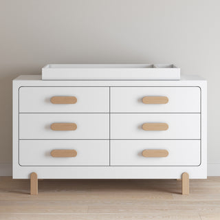 The Child Craft Legacy Changing Table Topper in Matte White sits atop a modern six-drawer white dresser with rounded wooden handles, providing a convenient diaper changing surface against a light beige wall and light wood floor.