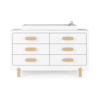 A modern Child Craft Legacy Changing Table Topper in matte white sits on a white double dresser with six drawers, light wood handles and legs, paired with a gray pad and pink bottle for a stylish, functional setup.