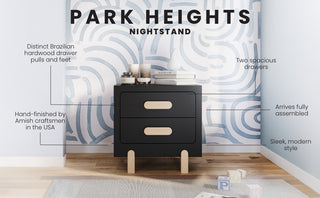 The Child Craft Park Heights Night Stand - Caviar Black features two spacious drawers with light wood pulls, crafted with Brazilian hardwood accents. It adds modern elegance to any room, especially against a bright, patterned blue wall.