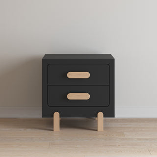 The Child Craft Park Heights Night Stand - Caviar Black is a modern two-drawer nightstand with light wood handles and Brazilian hardwood legs, elegantly placed against a white wall on a light wood floor.