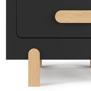 A close-up of the Child Craft Park Heights Night Stand - Caviar Black, a modern nightstand featuring a black cabinet, light Brazilian hardwood legs, and an oval wooden drawer handle.