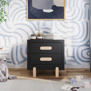 The Park Heights Night Stand in Caviar Black by Child Craft stands against a blue and white abstract wall, topped with decor, books, and candles. Children's books and a stuffed animal are placed on the wooden floor nearby.