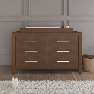 The Child Craft Kieran 6-Drawer Double Dresser in Toasted Chestnut sits against a gray paneled wall. A changing pad and baby essentials are on top, with a basket and plush toy nearby. A white patterned rug lies in front.