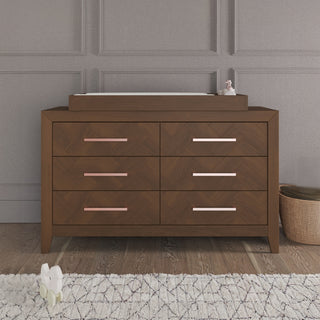 A Child Craft F01792 Universal Dressing Kit in Toasted Chestnut sits against a gray paneled wall, featuring six drawers with pink handles and a changing topper. Nearby, a woven basket and plush toy rest on the hardwood floor beside a patterned rug.