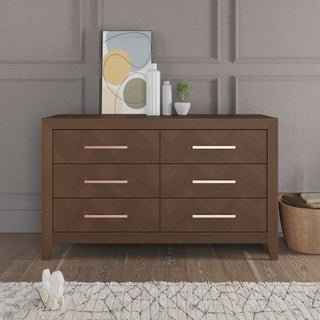 The Child Craft Kieran 6-Drawer Double Dresser in Toasted Chestnut stands against a paneled wall, topped with abstract art, two vases, and a small plant. A basket and plush toy are placed on the floor nearby.