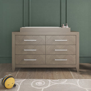 The Child Craft Kieran 6-Drawer Double Dresser in Crescent Gray stands against a green wall, topped with small containers and a cushioned changing pad. A plush bee toy rests on a patterned rug in the foreground.