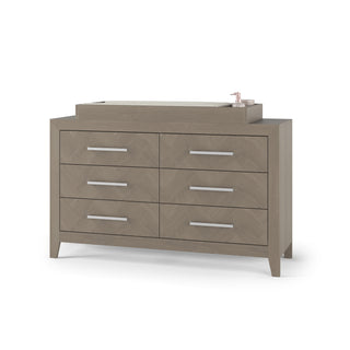 The Child Craft F01792 Universal Dressing Kit in Crescent Grey is shown on a dresser with modern metal handles, featuring a changing topper holding a pink bottle and baby essentials, all set against a white background.