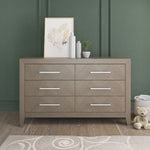 The Child Craft Kieran 6-Drawer Double Dresser in Crescent Gray stands against a green paneled wall, topped with framed art, bottles, and a plant. A plush teddy bear sits on the floor beside the dresser.