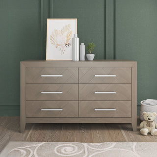 The Child Craft Kieran 6-Drawer Double Dresser in Crescent Gray stands against a green paneled wall, topped with framed art, bottles, and a plant. A plush teddy bear sits on the floor beside the dresser.