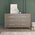 The Child Craft Kieran 6-Drawer Double Dresser in Crescent Gray stands against a green paneled wall, topped with framed art, bottles, and a plant. A plush teddy bear sits on the floor beside the dresser.