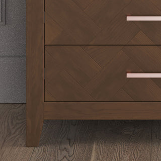 Close-up of the Kieran 5-Drawer Chest by Child Craft in Toasted Chestnut, showing its herringbone pattern, horizontal metal handles, and wooden legs on a wooden floor beside a gray textured wall.
